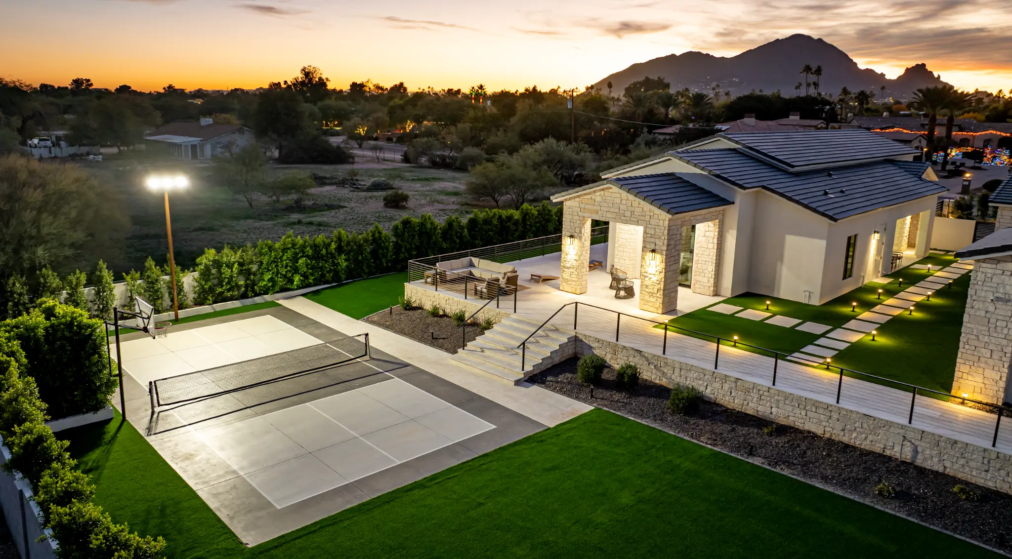 Aerial view of modern desert estate with pickleball court, stone columns, and illuminated grounds at dusk