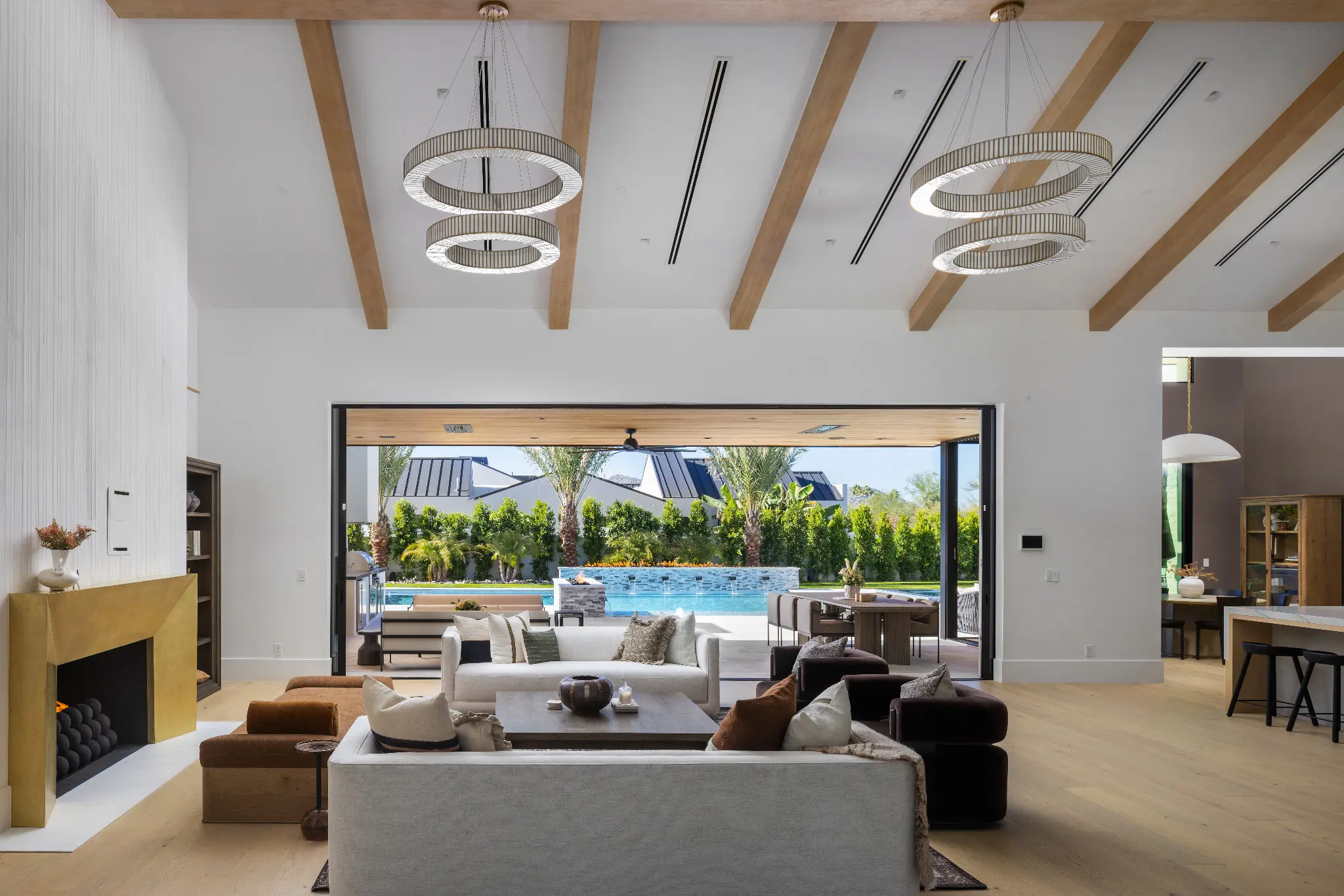Open-plan living room with exposed beam ceiling, ring chandeliers, and full-width pocket doors to pool